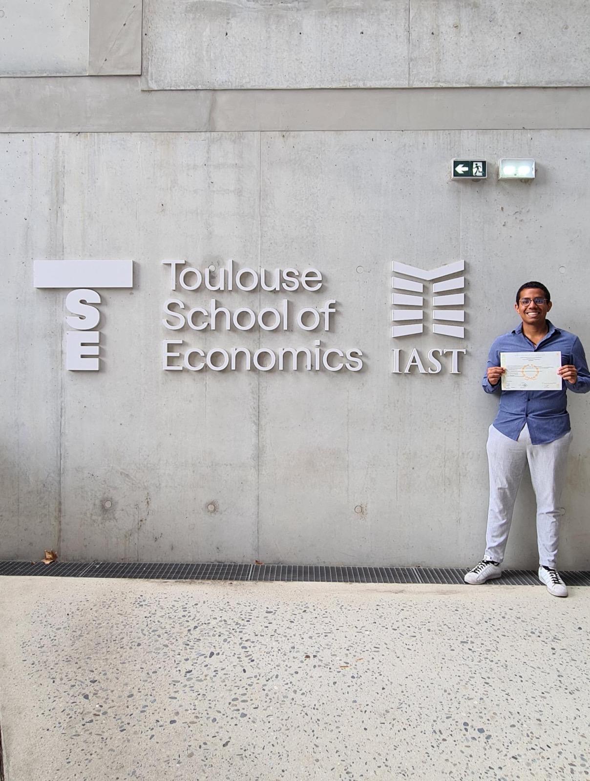Juan holding his economics diploma outside the Toulouse School of Economics